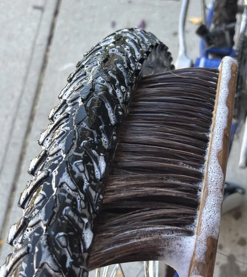 A soapy knobby tire being scrubbed with a wooden-handled bristle brush.